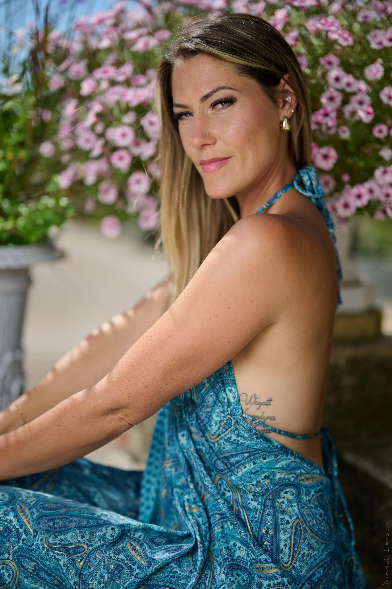 Femme souriante portant un vêtement bleu avec un motif paisley, assise devant un fond de fleurs roses, avec des boucles d'oreilles en or et un piercing au nez.