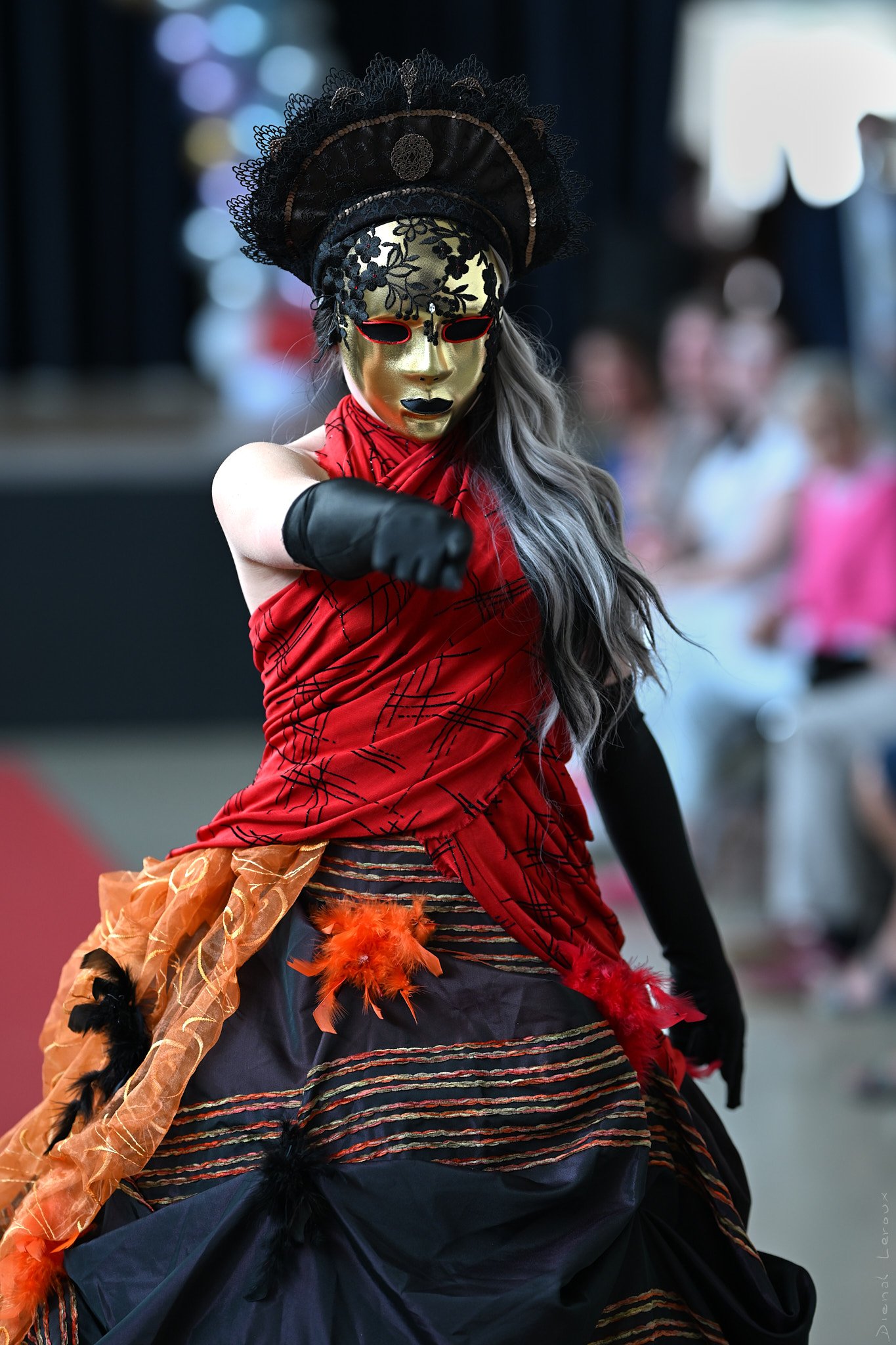 Une personne déguisée en carnaval portant un masque en or, un chapeau noir en dentelle, des vêtements rouges, noirs et orange avec des plumes, et des accessoires colorés. La personne fait un geste avec le bras tendu. L'arrière-plan est flou avec d'autres personnes présentes.