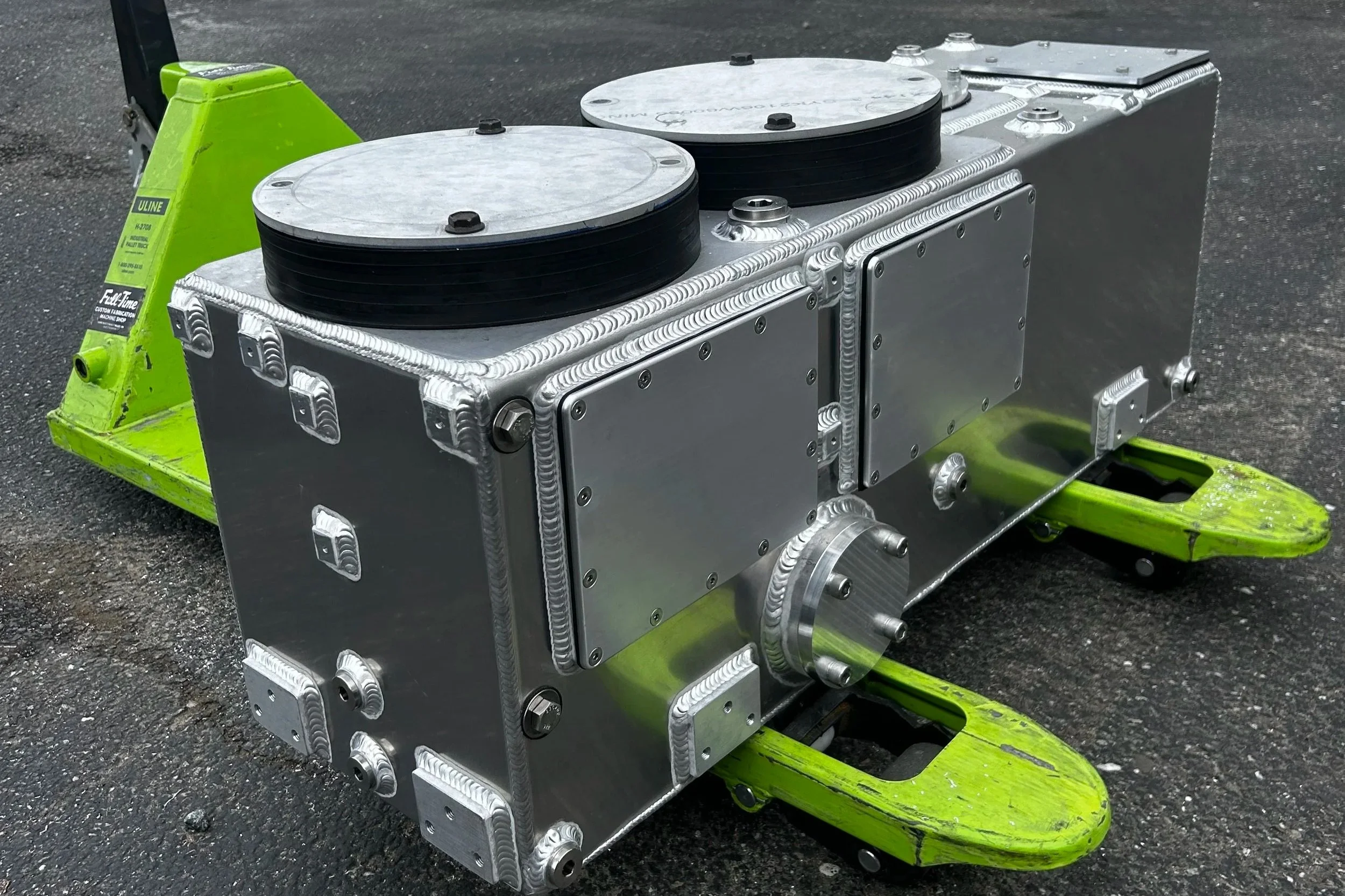 Close-up of a metallic robot component with black circular elements on top, mounted on a green dolly on a paved surface.
