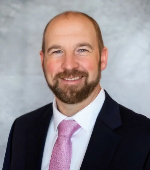A man with a beard and short hair wearing a dark suit, white shirt, and pink tie, smiling against a gray background.