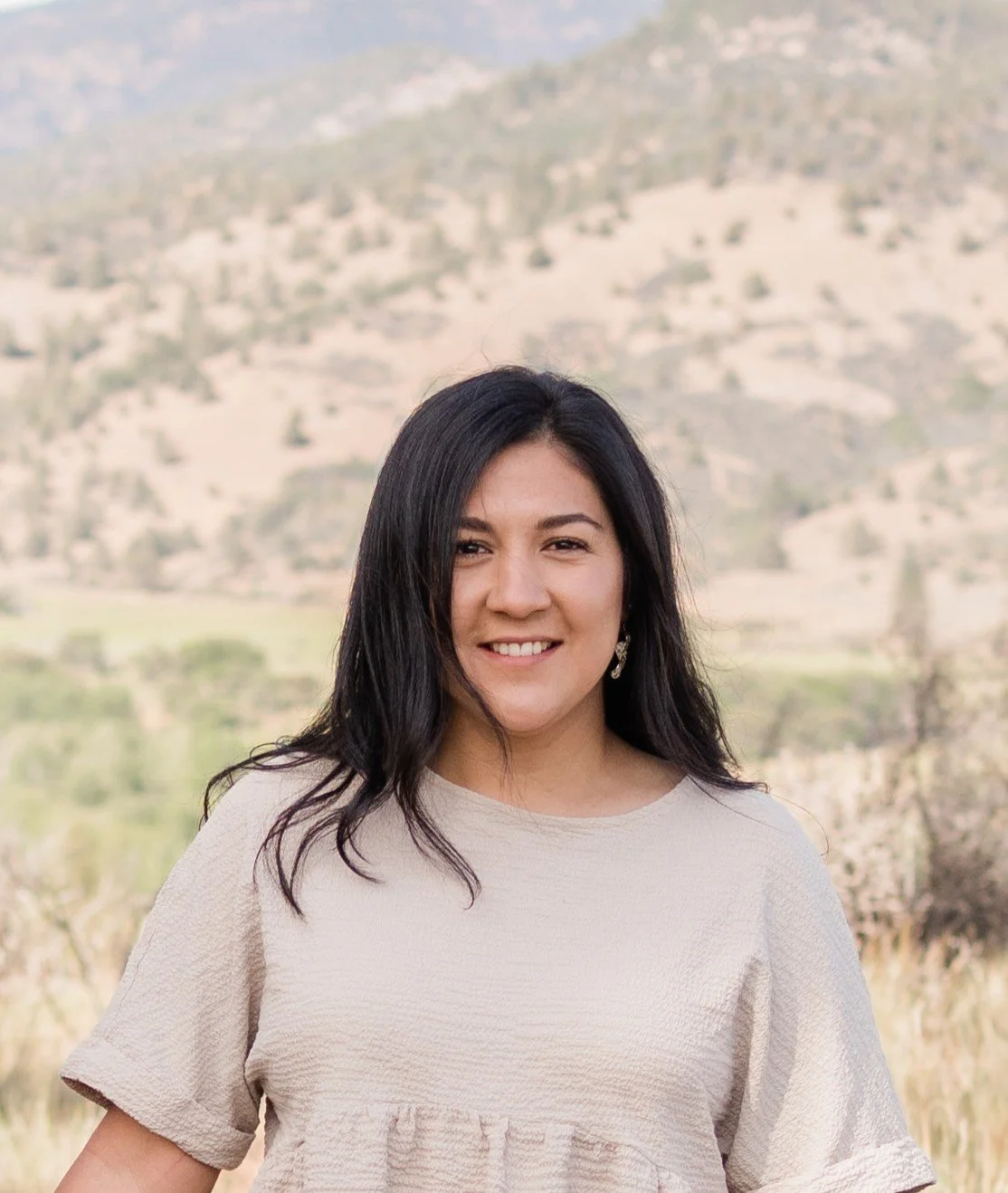 A woman with dark hair wearing a beige top standing outdoors with a mountainous landscape in the background.