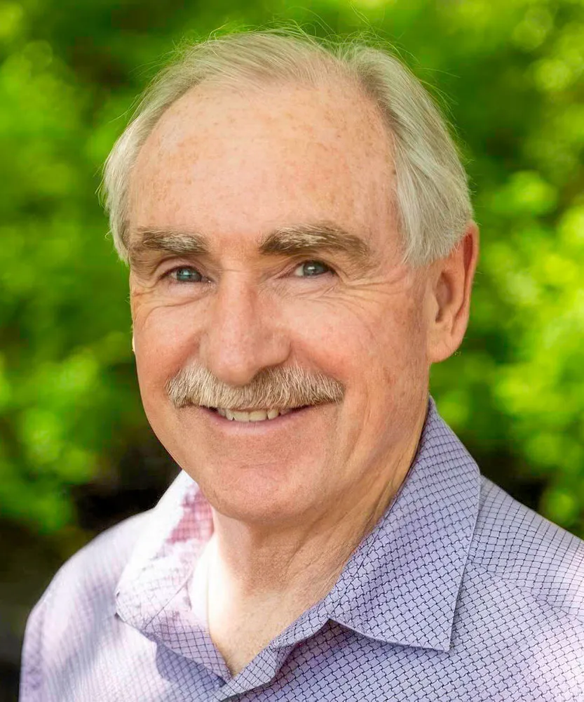 Close-up portrait of smiling man with gray hair and mustache, wearing a light-colored, patterned shirt, outdoors with green blurred foliage in the background.