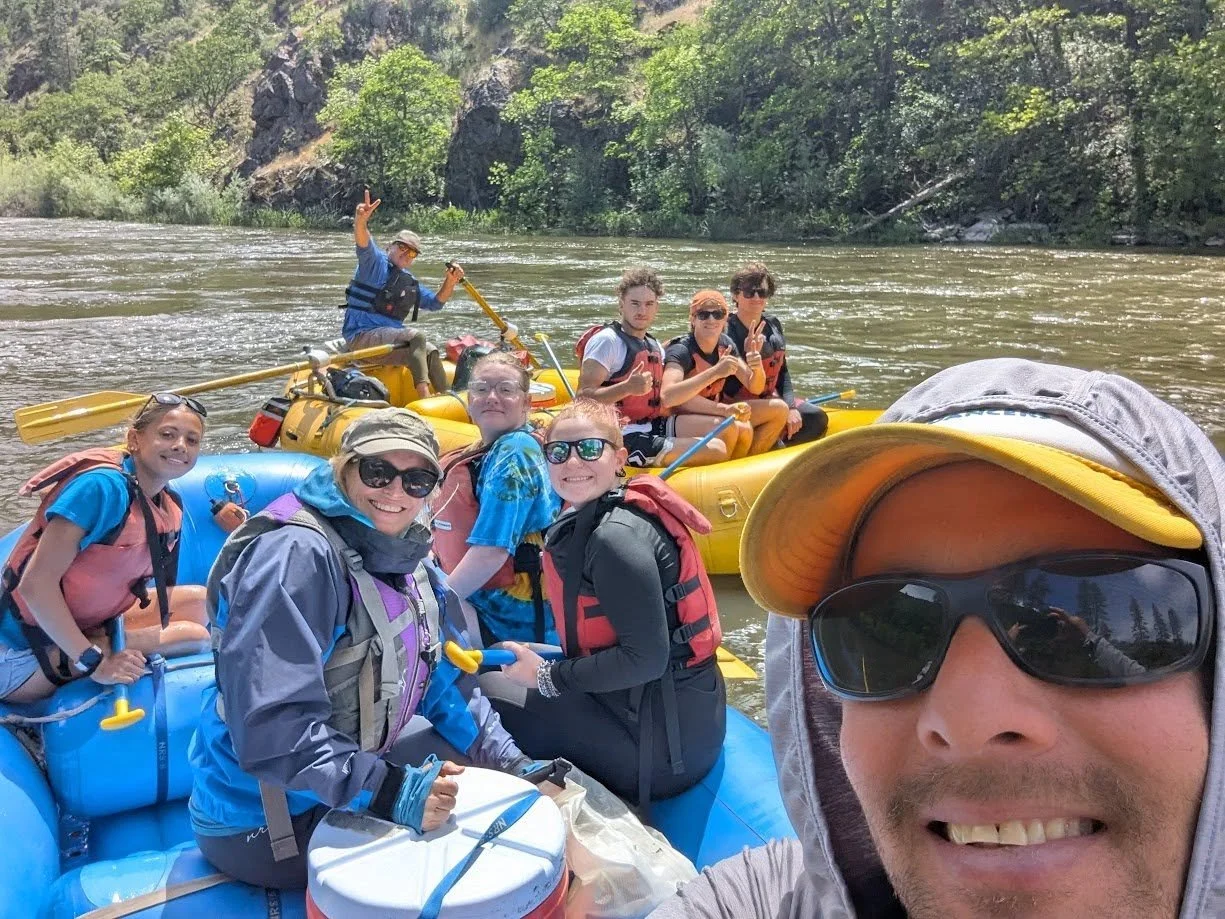 Group of people on two inflatable rafts on a river, surrounded by trees and rocky hills, with one person in the foreground taking a selfie and the others smiling and making peace signs.