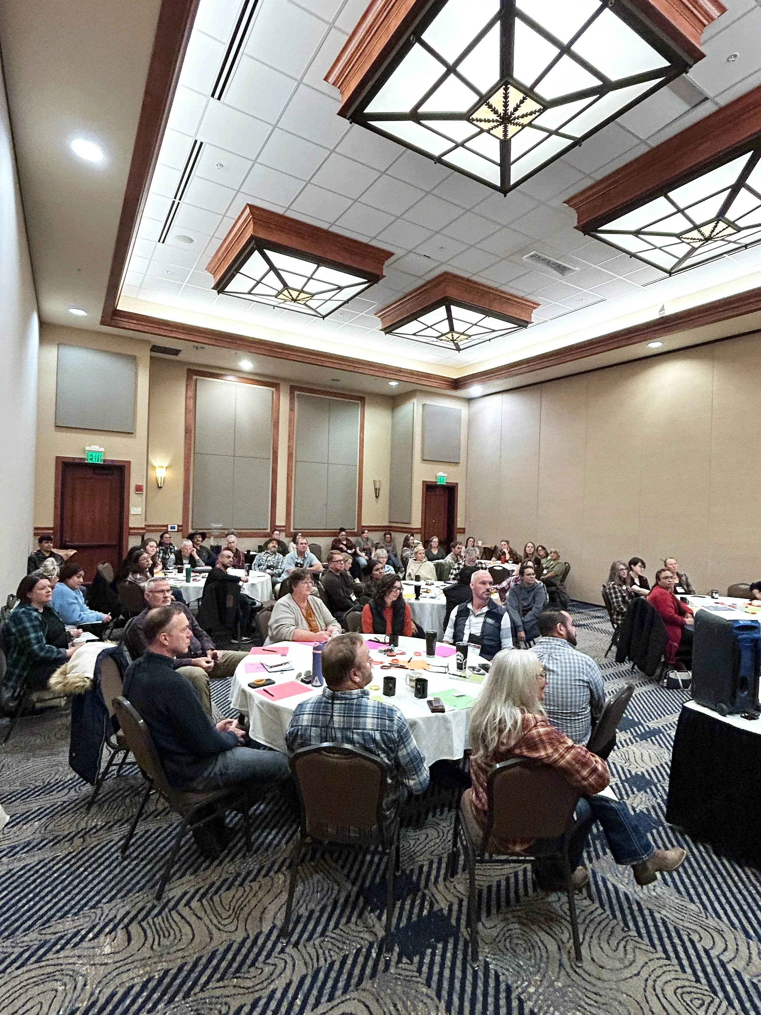 A large conference room filled with people sitting at round tables, listening attentively during a presentation or seminar. The room has a high ceiling with decorative lighting fixtures and wood paneling.