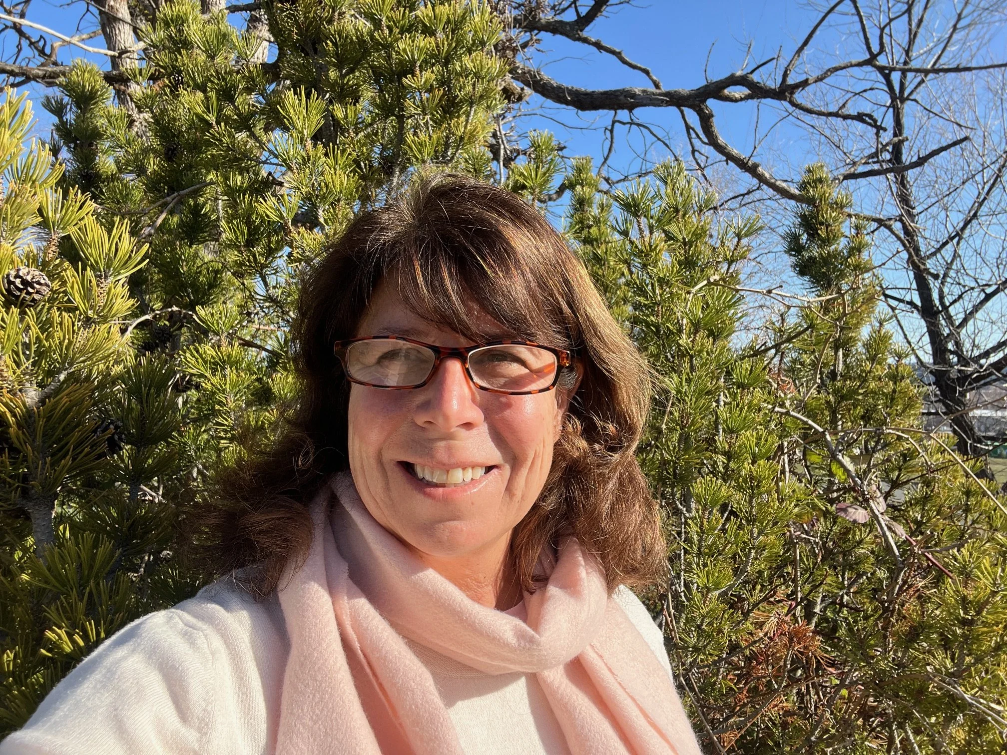 A woman with glasses smiling outdoors in front of green shrubs and trees on a sunny day.