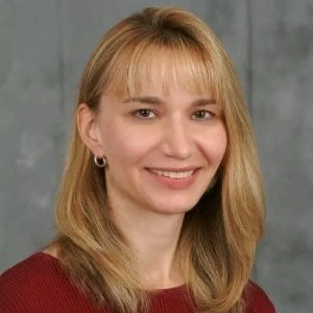 A woman with blonde hair, wearing a red top, smiling, and standing against a gray background.