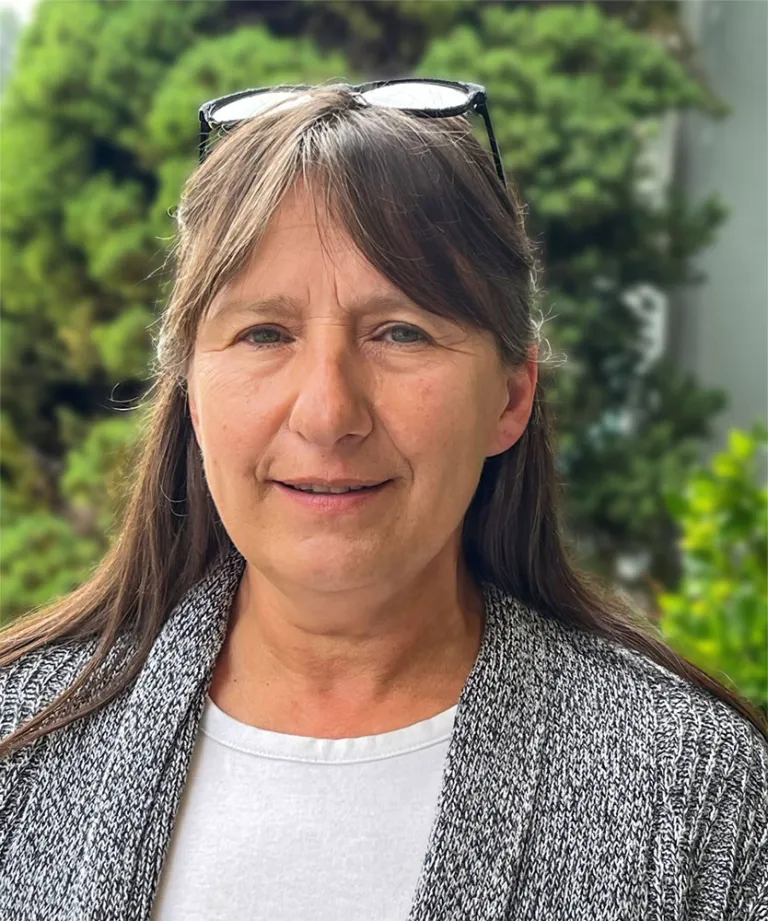 Close-up of a middle-aged woman with long brown hair, wearing glasses on top of her head, a white shirt, and a gray patterned jacket, outdoors with green trees in the background.