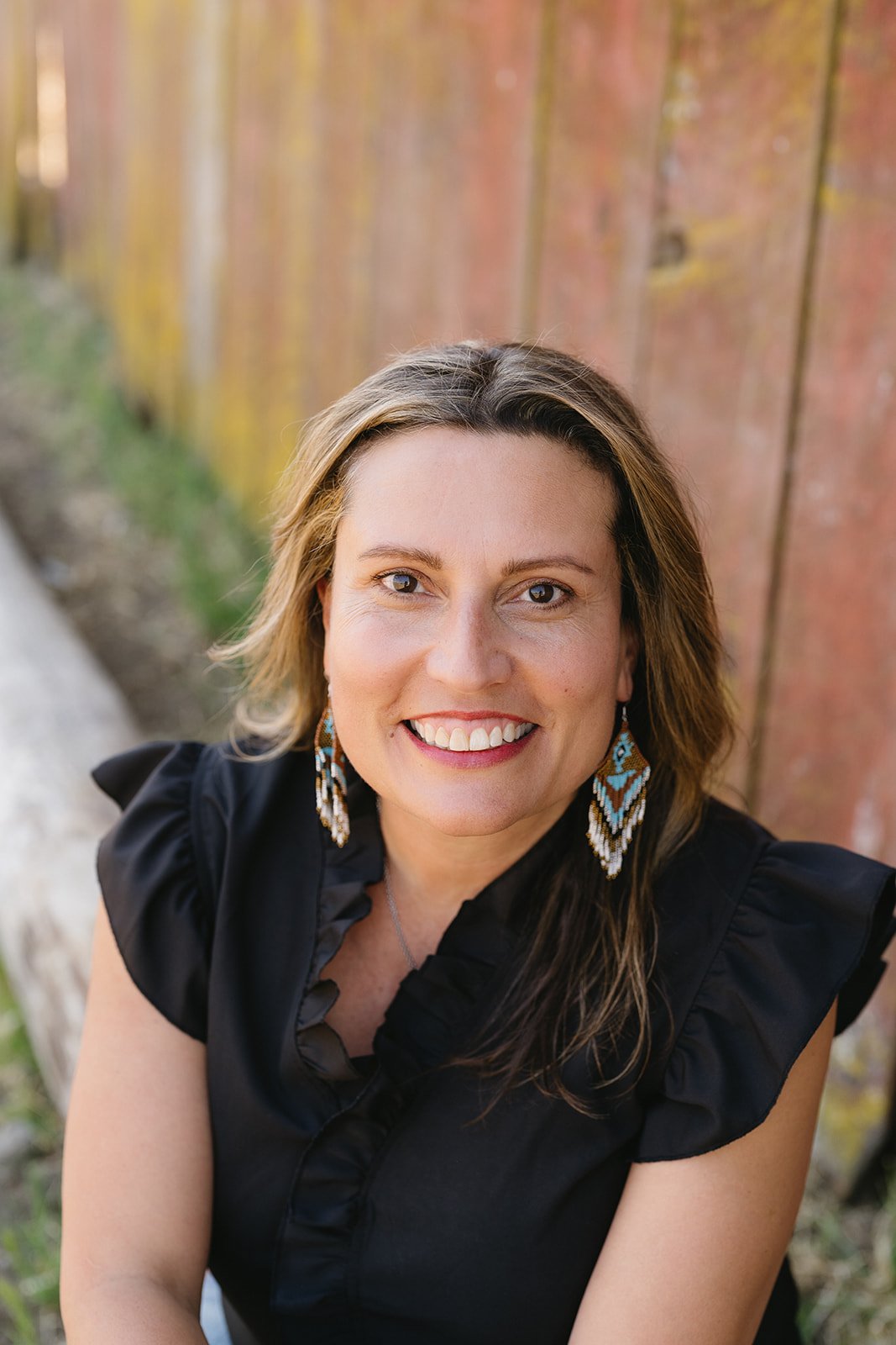 A woman with shoulder-length brown hair, wearing earrings with blue, white, and brown diamond patterns and a black ruffled top, sitting outdoors against a wooden fence, smiling at the camera.