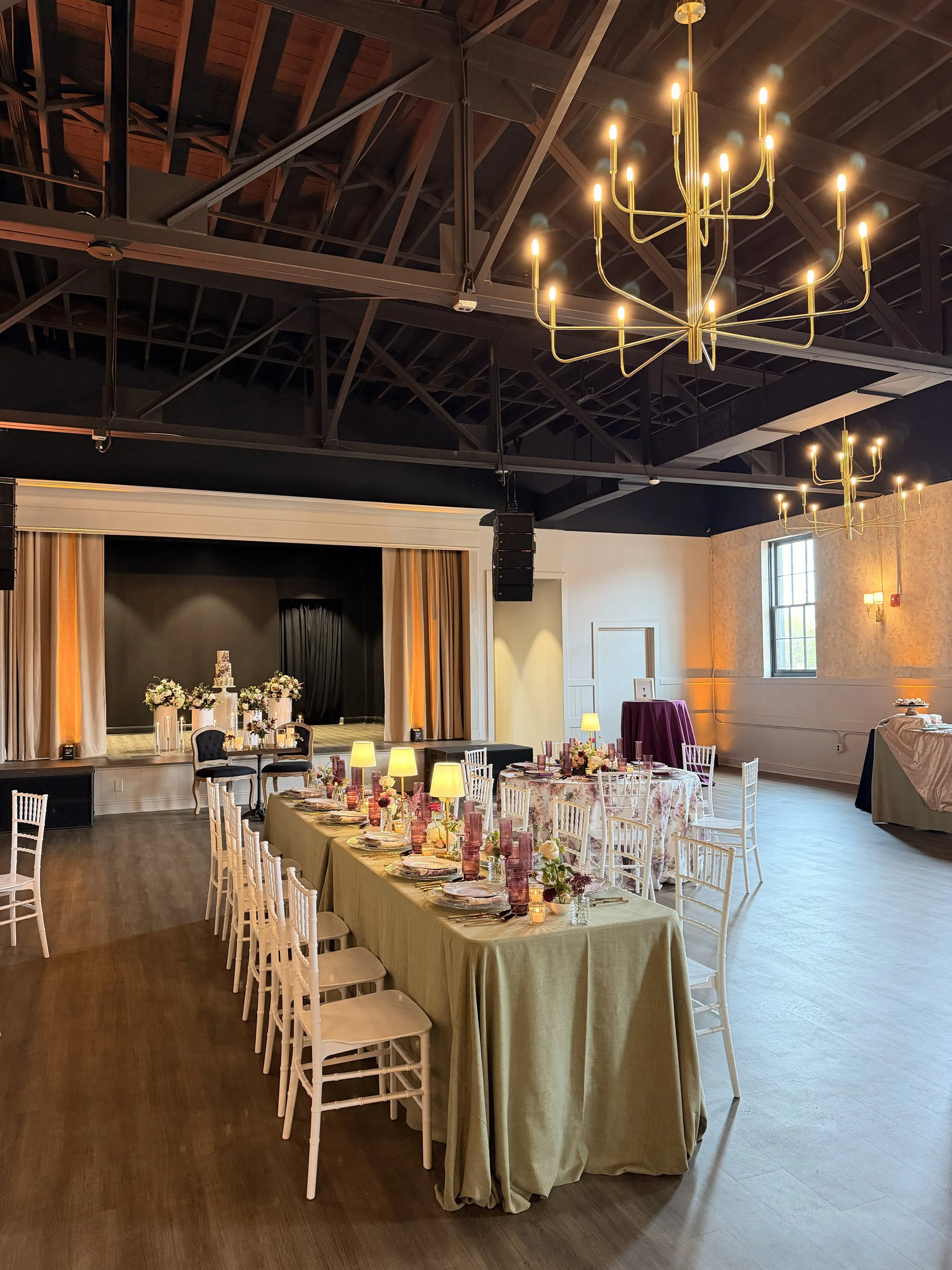 A long dining table with white tablecloths, white folded napkins, and silverware in a bright, rustic restaurant with wooden beams, large windows, and string lights overhead.