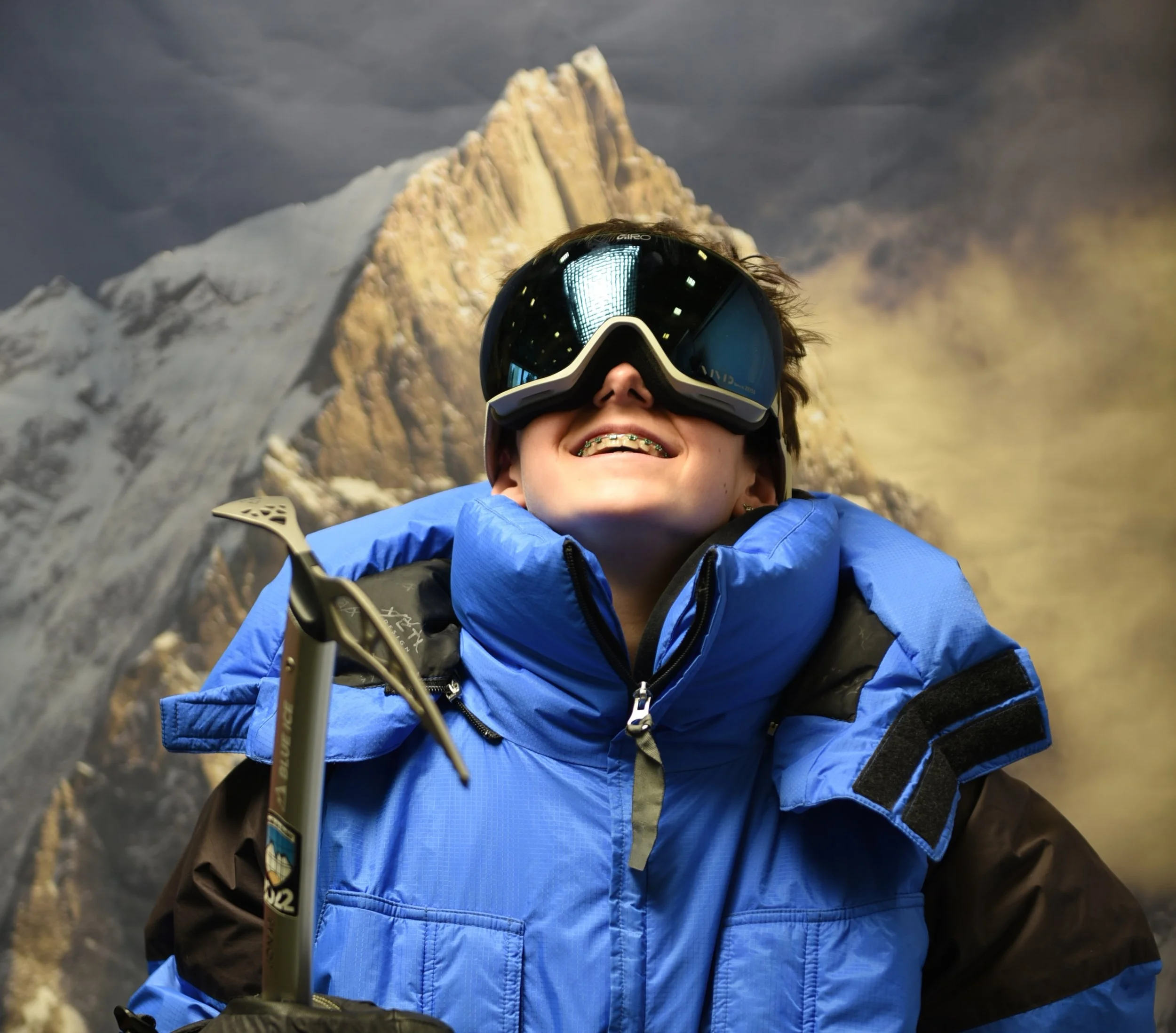 Skifahrer auf einem schneebedeckten Berggipfel mit Blick auf schneebedeckte Berge im Hintergrund, trägt einen Helm, eine blaue Jacke, eine orange Tasche und benutzt Skistöcke.