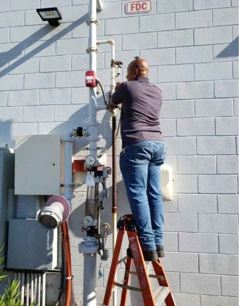 A person standing on a step ladder repairing pipes and gauges on an exterior wall marked with an 'FDC' sign.