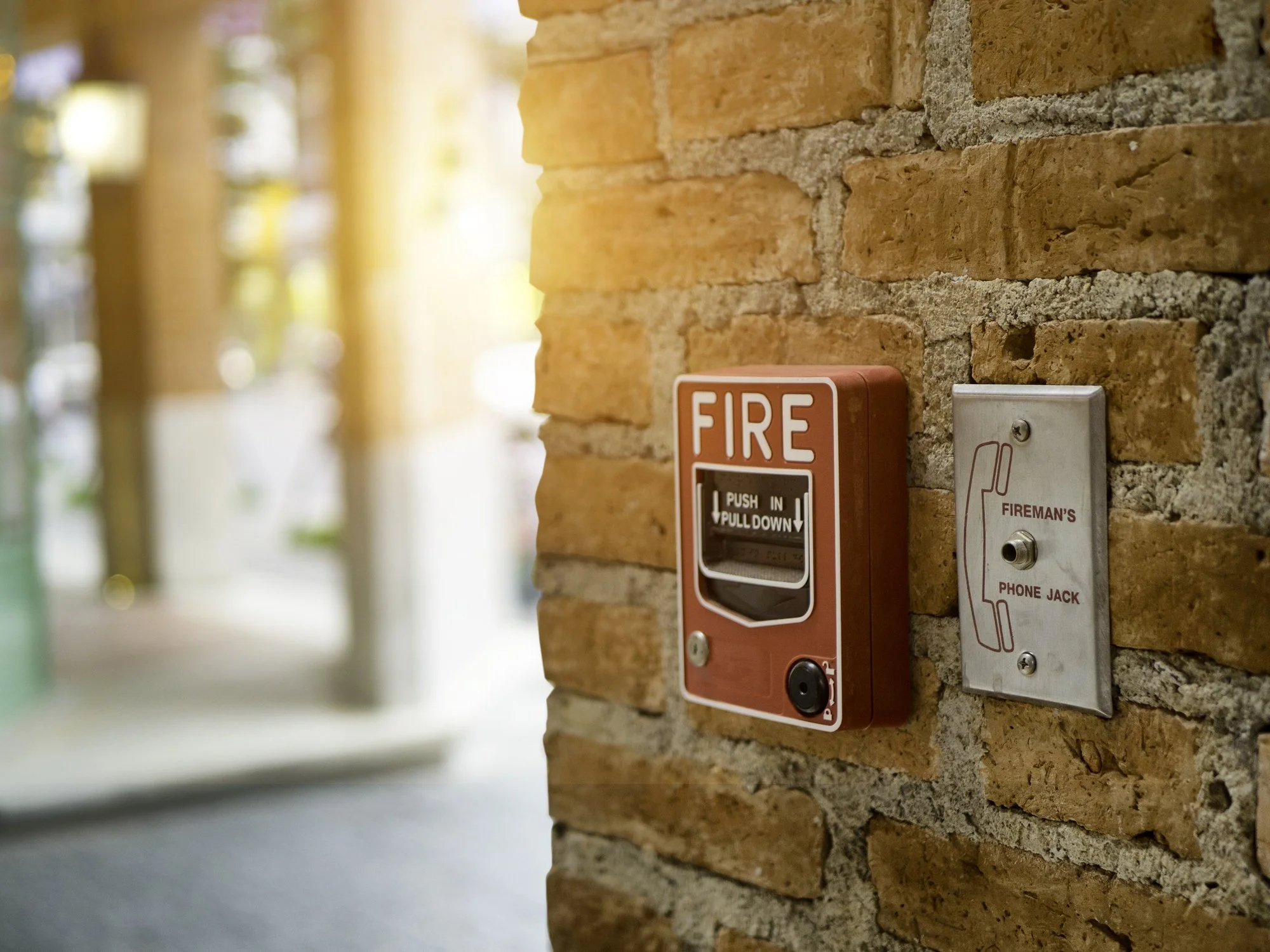 A red fire alarm box and a gray fireman's phone jack mounted on a brick wall in a hallway with sunlight streaming in.