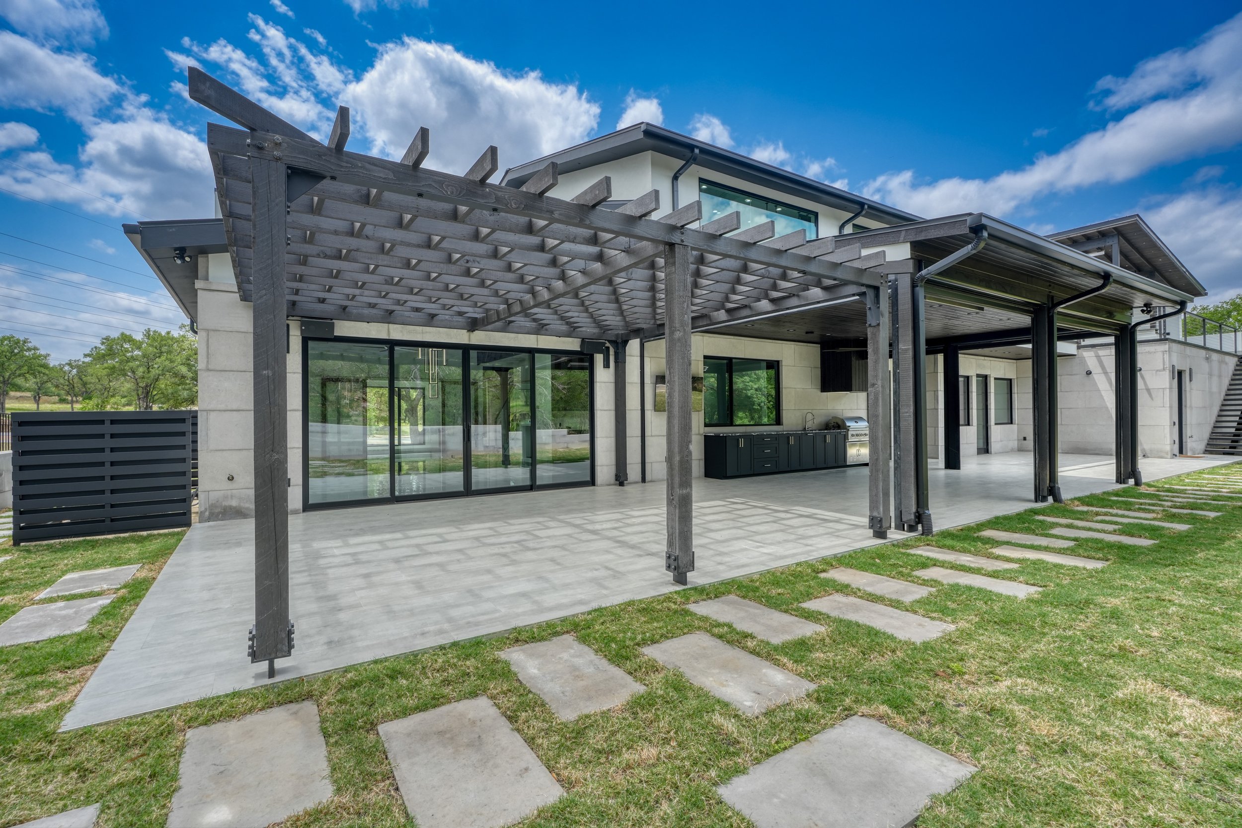 Modern house exterior with a covered patio area, outdoor kitchen, and a pergola, overlooking a grassy lawn with stone stepping paths and a blue sky with clouds.