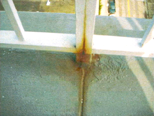 Corrosion and rust staining at a balcony railing post base on a coated deck.