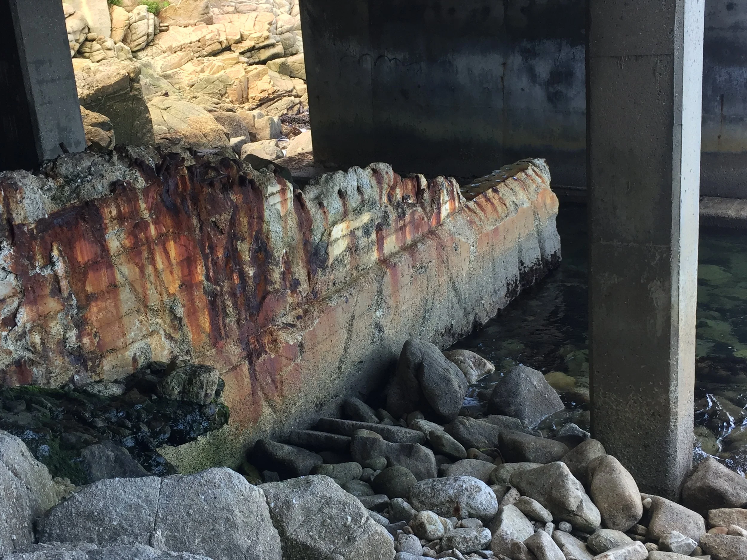 Severely deteriorated concrete structure beneath a coastal plaza, showing exposed and corroded reinforcement due to prolonged saltwater exposure.
