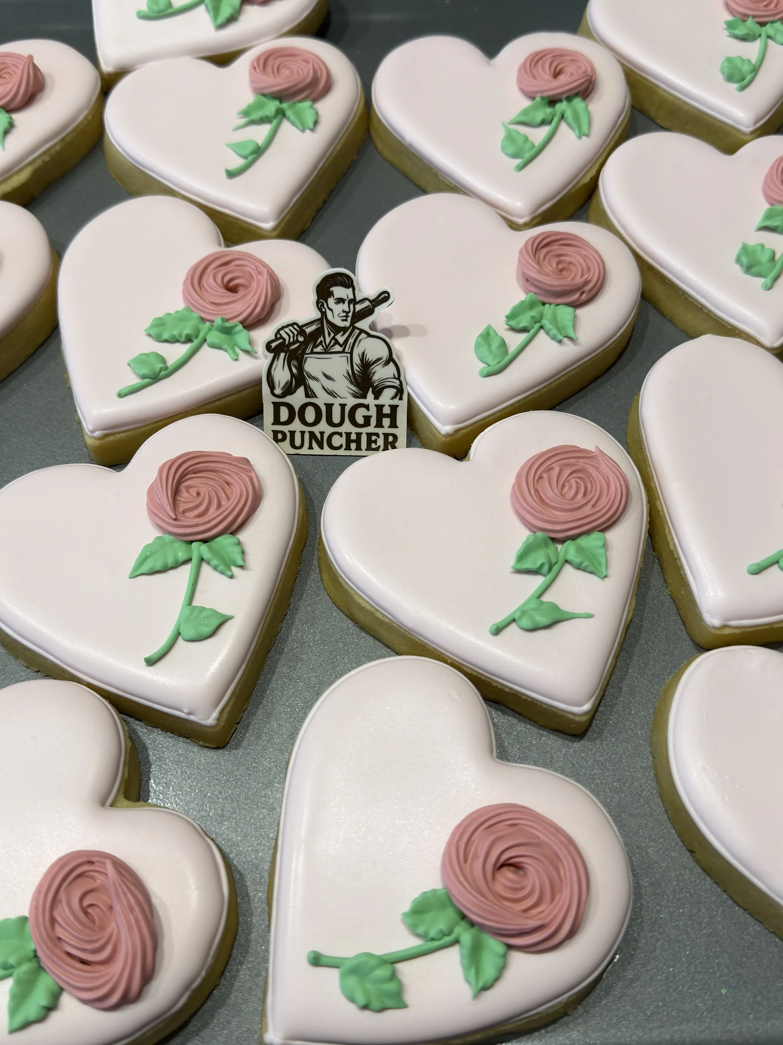 Heart-shaped iced sugar cookies decorated with pink frosting roses and green leaves, arranged around a Dough Puncher logo.