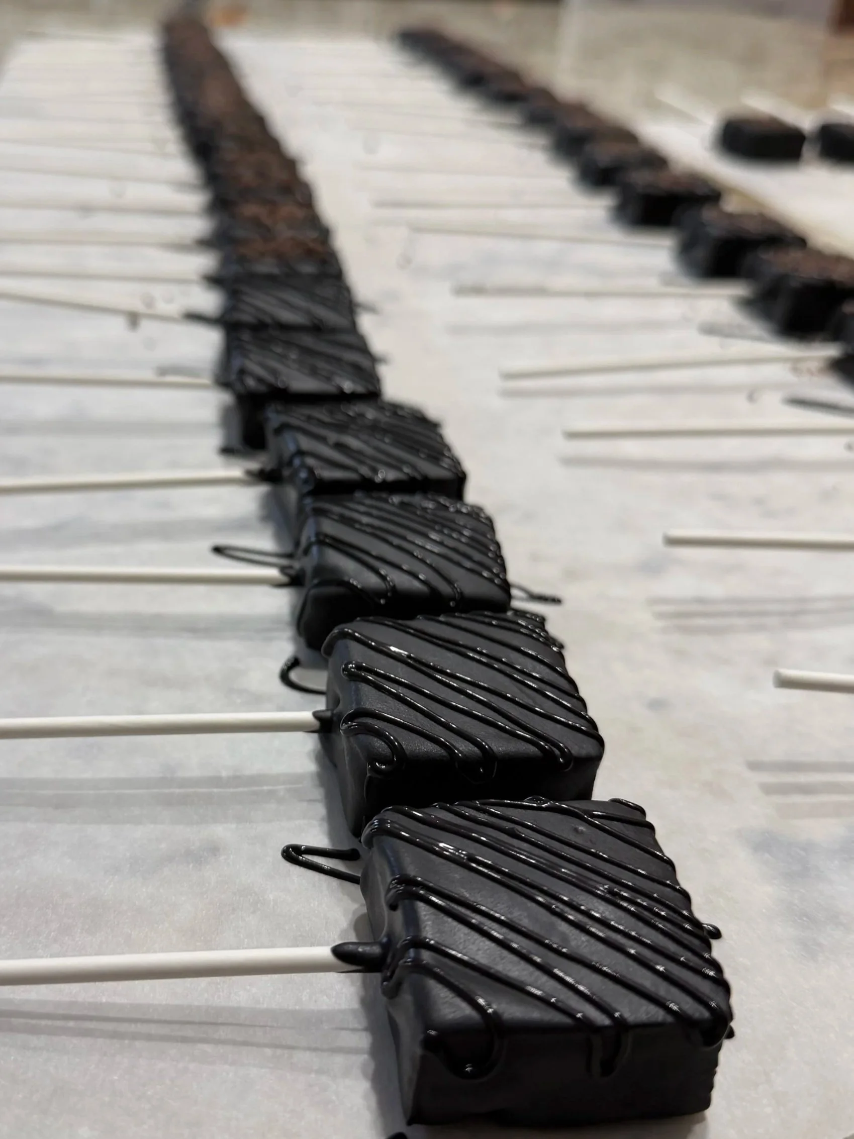 Rows of black chocolate-coated cake pops on white sticks cooling on a tray