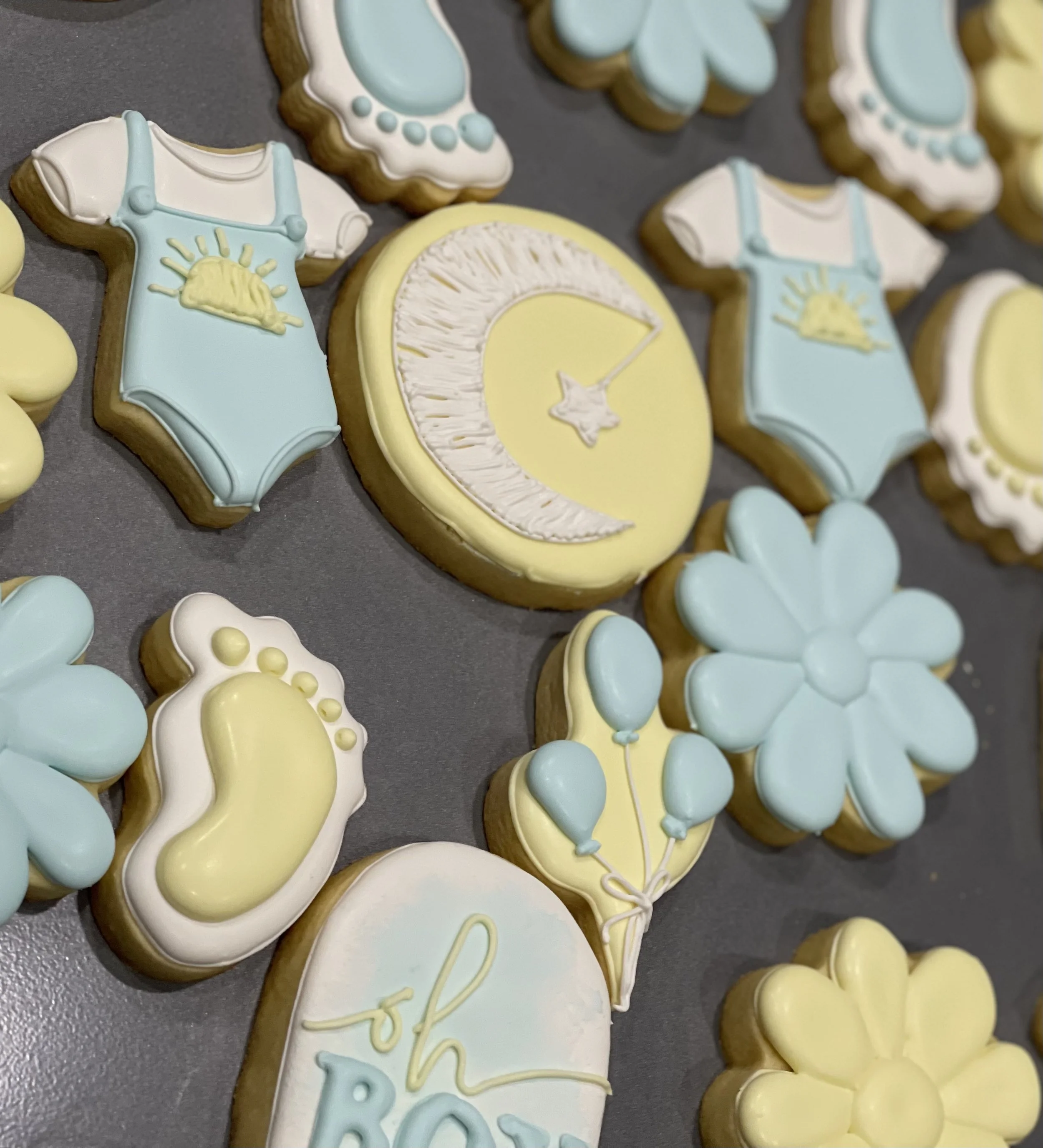 Close-up of pastel blue and yellow decorated baby shower cookies, including onesies, baby feet, balloons, flowers, and a moon-and-star design.