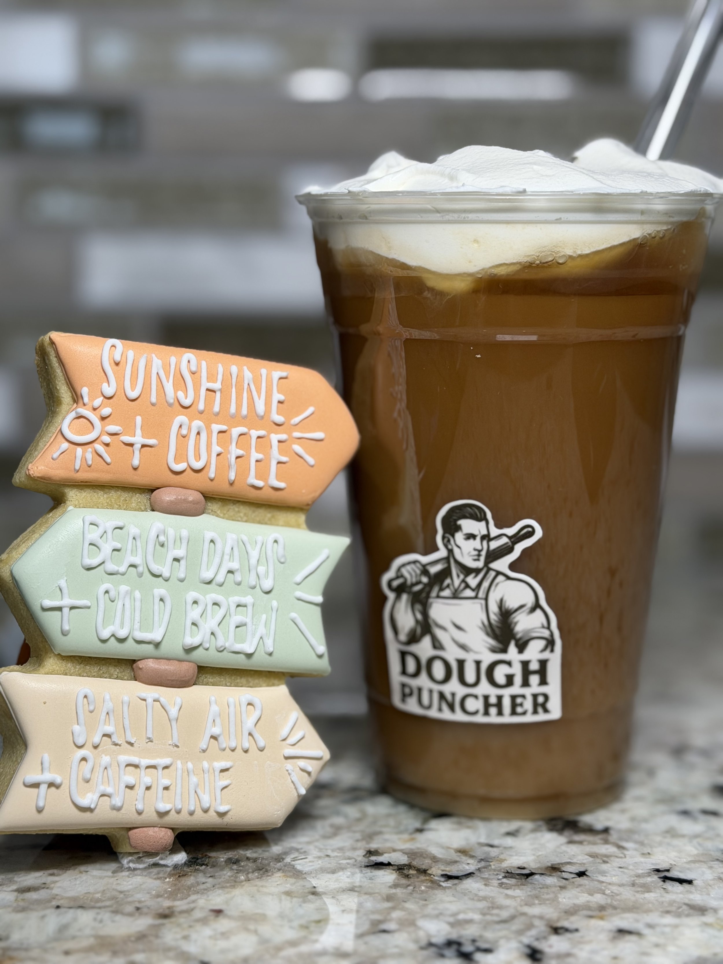 Glass of iced coffee with whipped cream beside colorful motivational cookies and a Dough Puncher sticker