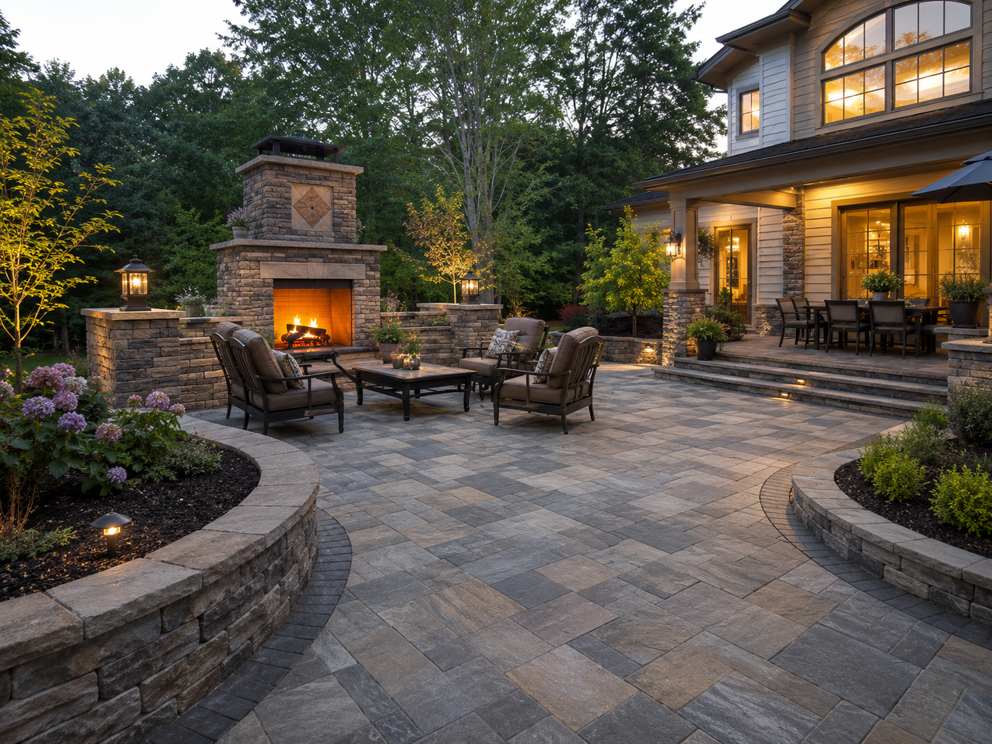 Outdoor paver patio with stone paving, a fireplace, and seating area at dusk, with a house in the background and trees surrounding the yard.