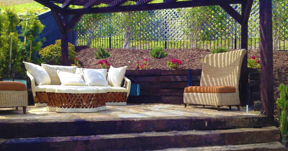 Outdoor patio with white cushioned bench and beige high-back chair under a pergola, landscaped garden with pink flowers in background.