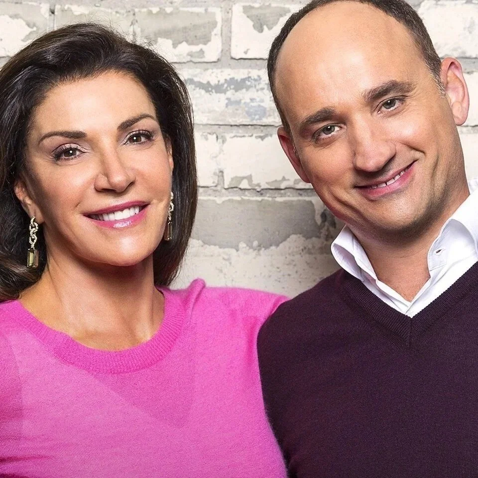 A woman and a man smiling, standing close together, with a white brick wall in the background.