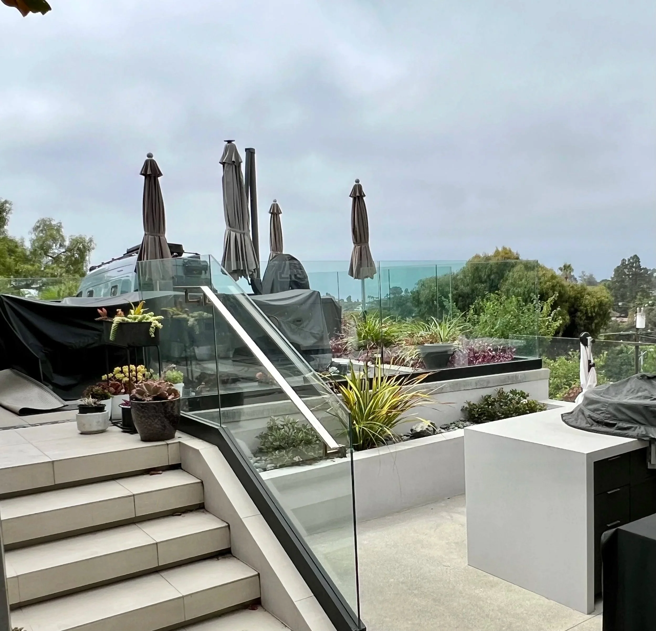 Outdoor terrace with potted plants, steps, umbrellas, and glass railing, overlooking greenery and cloudy sky.