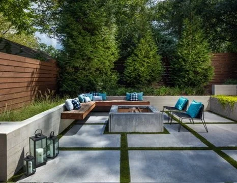 Modern backyard patio with concrete pavers, a wooden L-shaped bench with blue and white pillows, two blue chairs, a concrete fire pit in the center, and decorative lanterns, surrounded by greenery and trees.