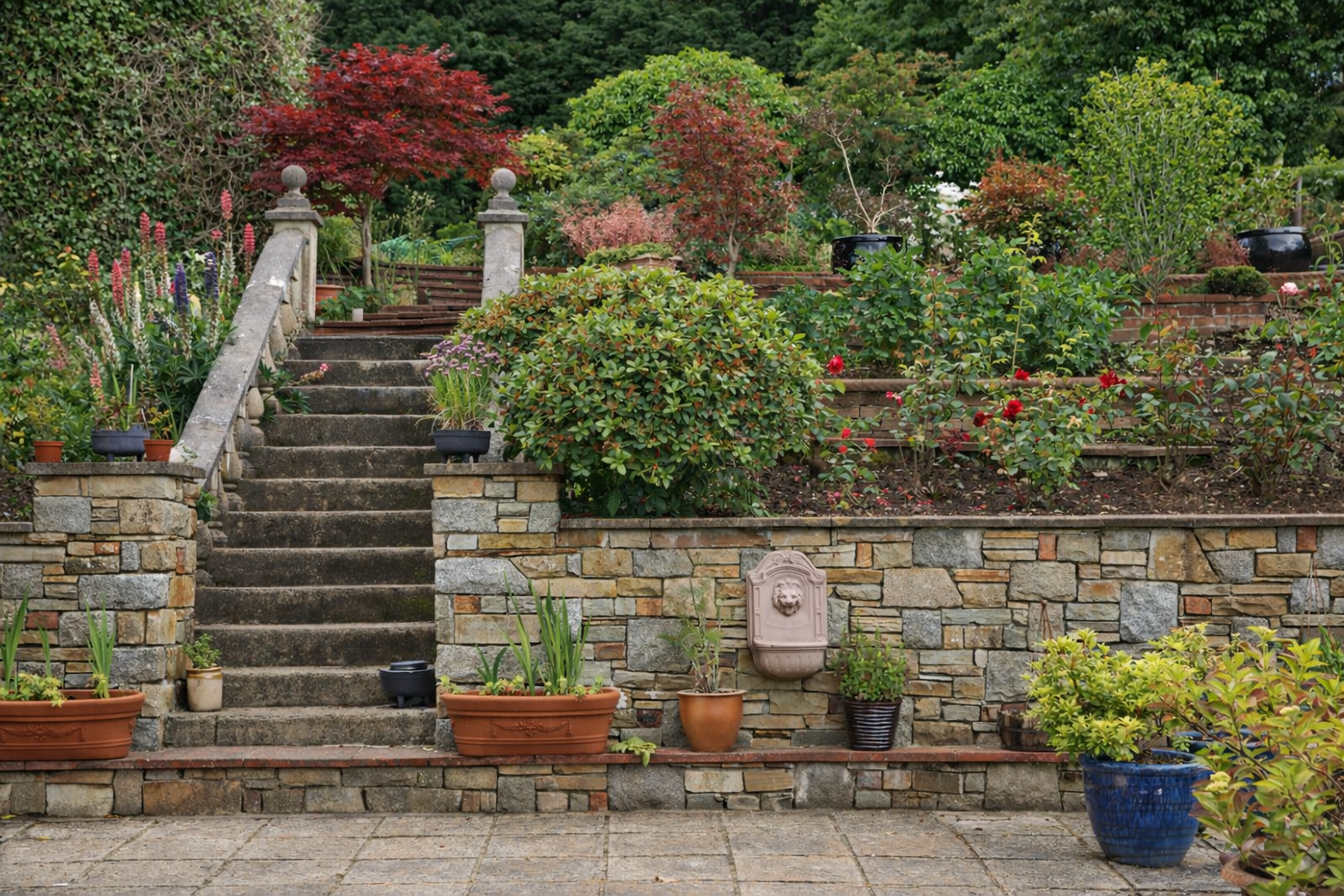 A garden staircase with stone steps leading up a hill, bordered by a stone wall with potted plants and a decorative fountain, surrounded by lush greenery, flowering bushes, and trees.