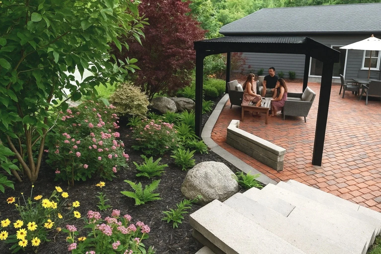 3D Design, A backyard patio with a brick floor, a covered seating area with four people sitting and talking, surrounded by lush green plants, flowers, rocks, and a house with gray siding and a patio table with an umbrella in the background.