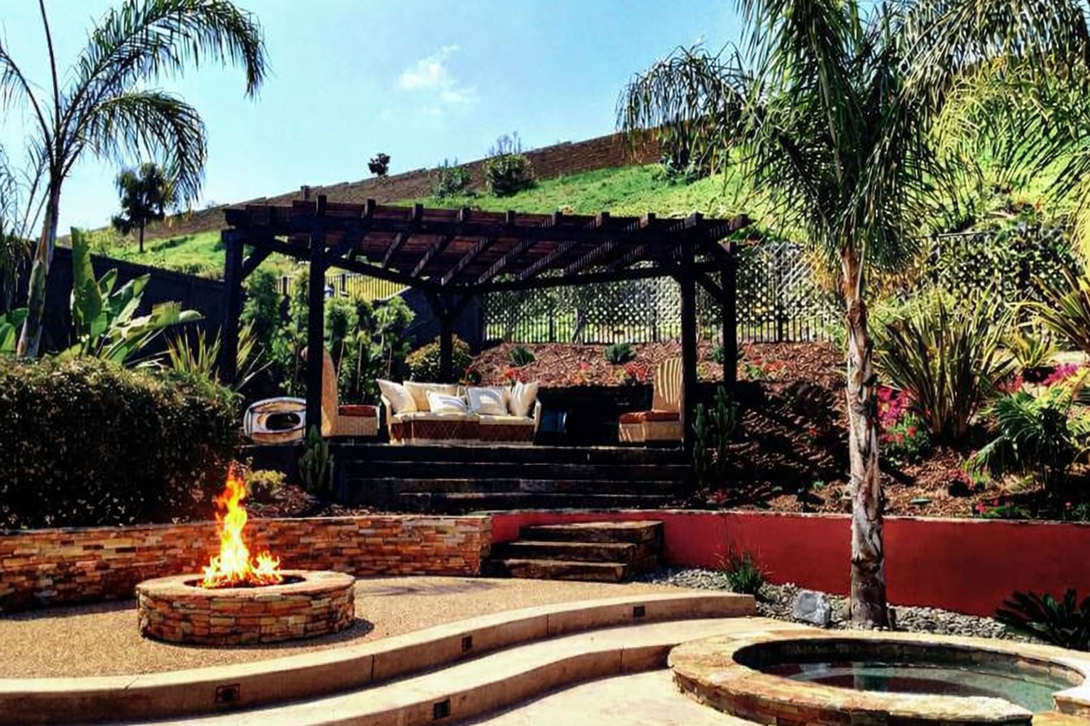 Backyard outdoor patio area with a fire pit, a covered seating area with cushioned outdoor furniture, and lush green plants and trees. Fire pit Contractor near me in San Diego.  Who is spa contractors near me?