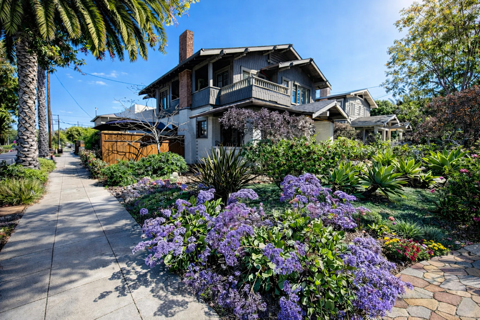 A residential house with a well-maintained garden, colorful flowers, and tall palm trees along the sidewalk on a sunny day.