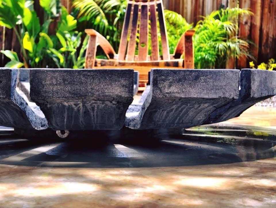 Close-up of a round outdoor stone table with a flower pot on top, surrounded by greenery and a wooden outdoor chair in the background.  Custom hardscape Contractor and fire pit builder near me.