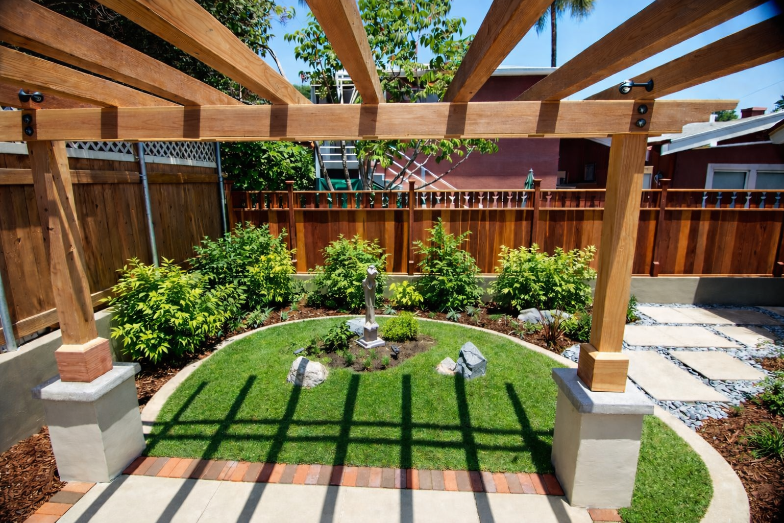 View of a backyard garden with a small lawn, a white statue, rocks, and green plants, surrounded by a wooden fence and a partially built wooden pergola.