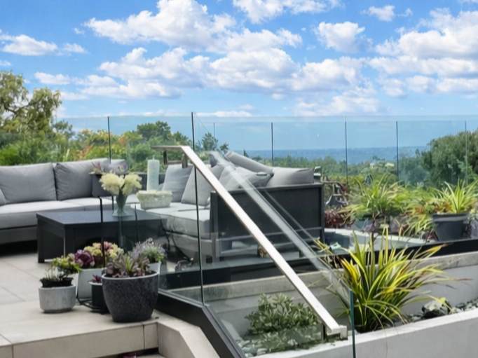 Patio outdoor living space with gray contemporary furniture, decorative potted plants, and a modern glass railing overlooking scenic tree-lined views in La Jolla