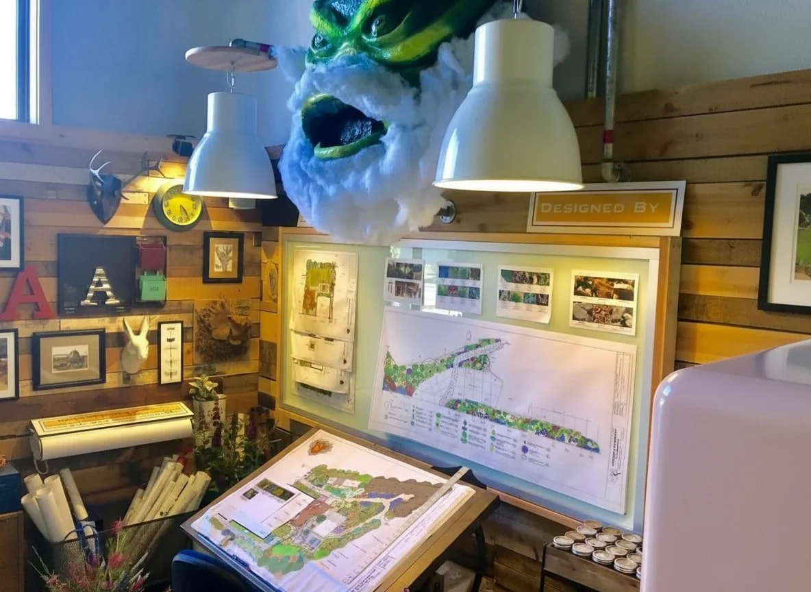 An art studio or design office with architectural drawings and visual projects on display. A large, decorative mask or sculpture with an angry face and green eyebrows hangs from the ceiling. Nature's Elements Landscaping