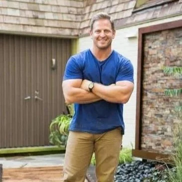 A smiling man standing outdoors with arms crossed, wearing a blue t-shirt and khaki pants.