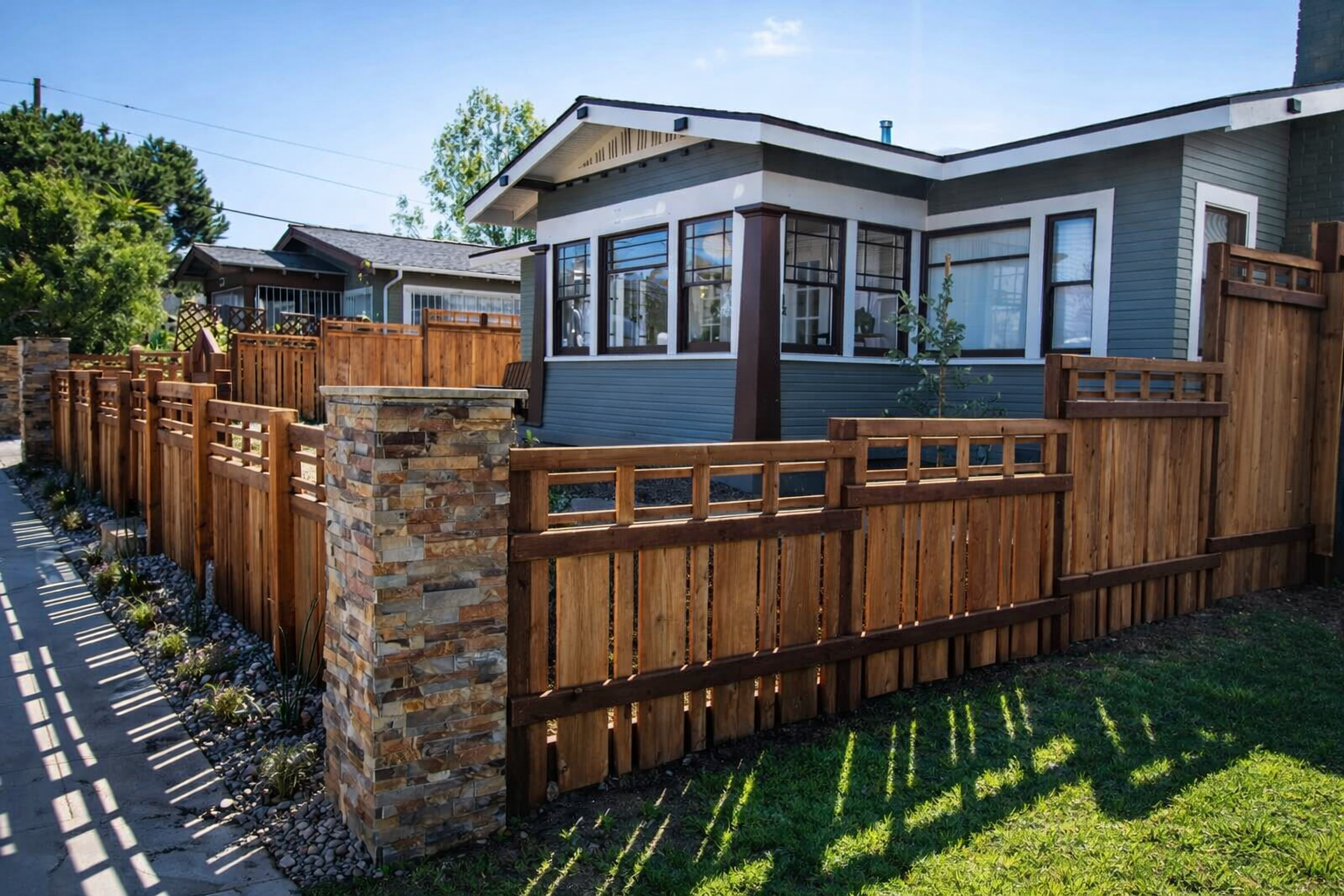 Residential backyard with a blue house, wooden fence, stone pillars, and a garden bed along the walkway, sunny day.  Landscape lighting contractor near me