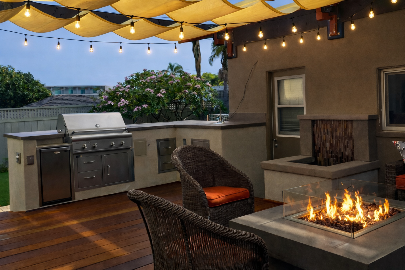 Outdoor patio with string lights, a built-in gas grill, wicker chairs with orange cushions, a fire pit, and a backyard garden with flowering bushes. Contractor, who can build an outdoor kitchen near me. 