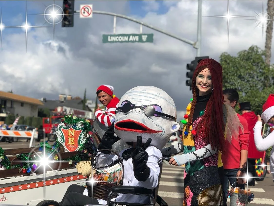 People participating in a Christmas parade, dressed in festive costumes, with one person in a large white mask with sunglasses, and a woman with red hair smiling, wearing a colorful outfit, standing next to them.  Nightmare before Christmas parade