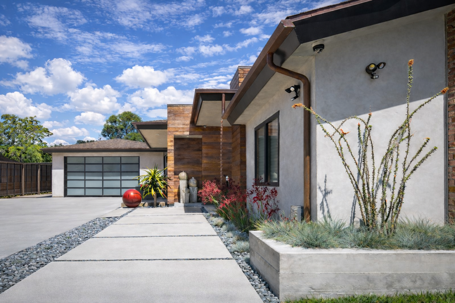 Modern house entrance in La Jolla with concrete walkway, drought-tolerant plants, and exterior lighting, under a partly cloudy blue sky. Custom Landscape who does Hardscape near me in La Jolla?