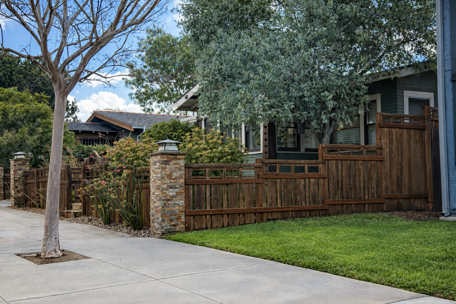 A residential house with a wooden fence, stone pillars, and green landscaping, including a large tree and bushes.