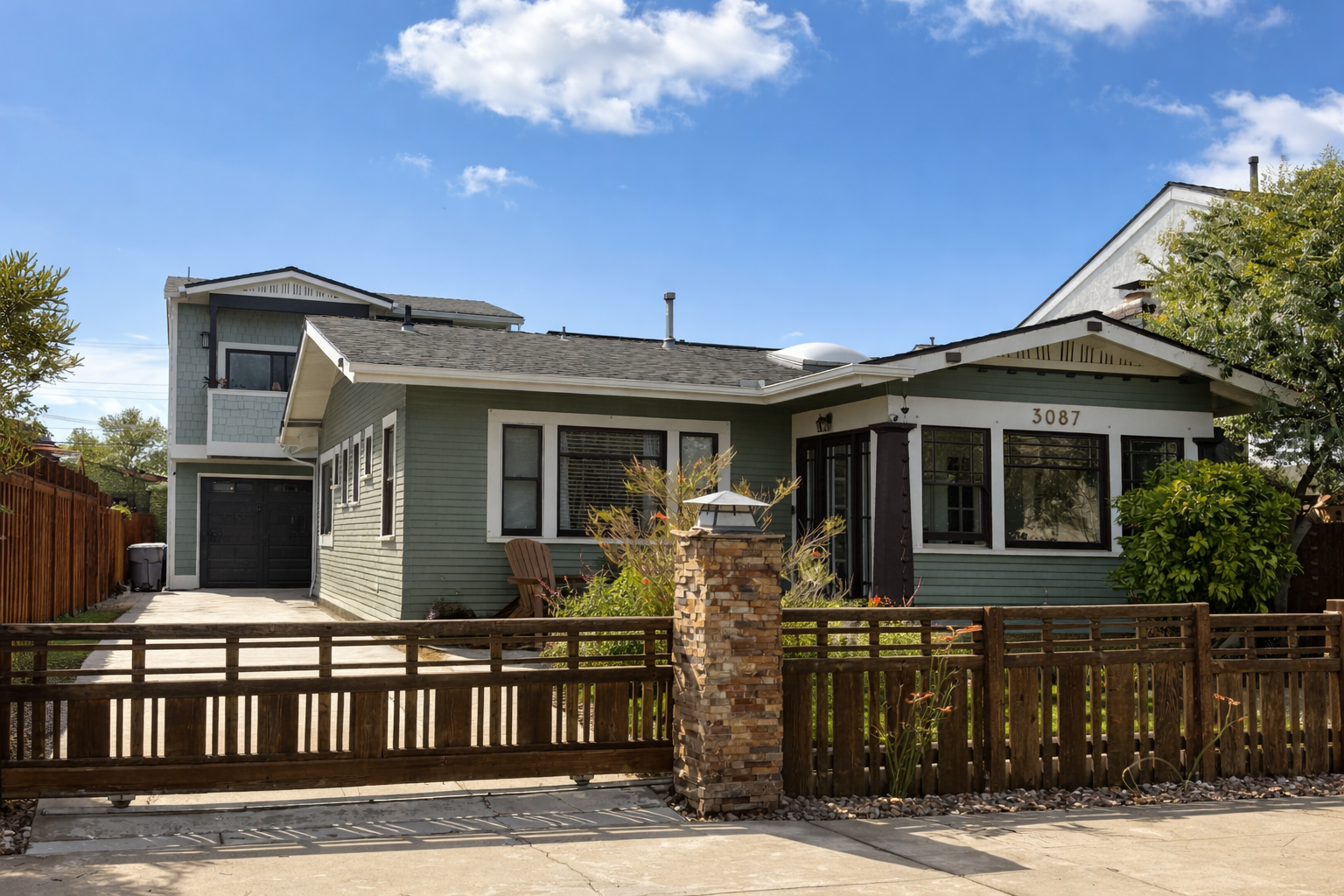A two-story house with a front yard, wooden fence, and a stone pillar with a lantern on top. The house has light green siding, black window frames, and a black garage door. There are plants and shrubs in the front yard under a blue sky.