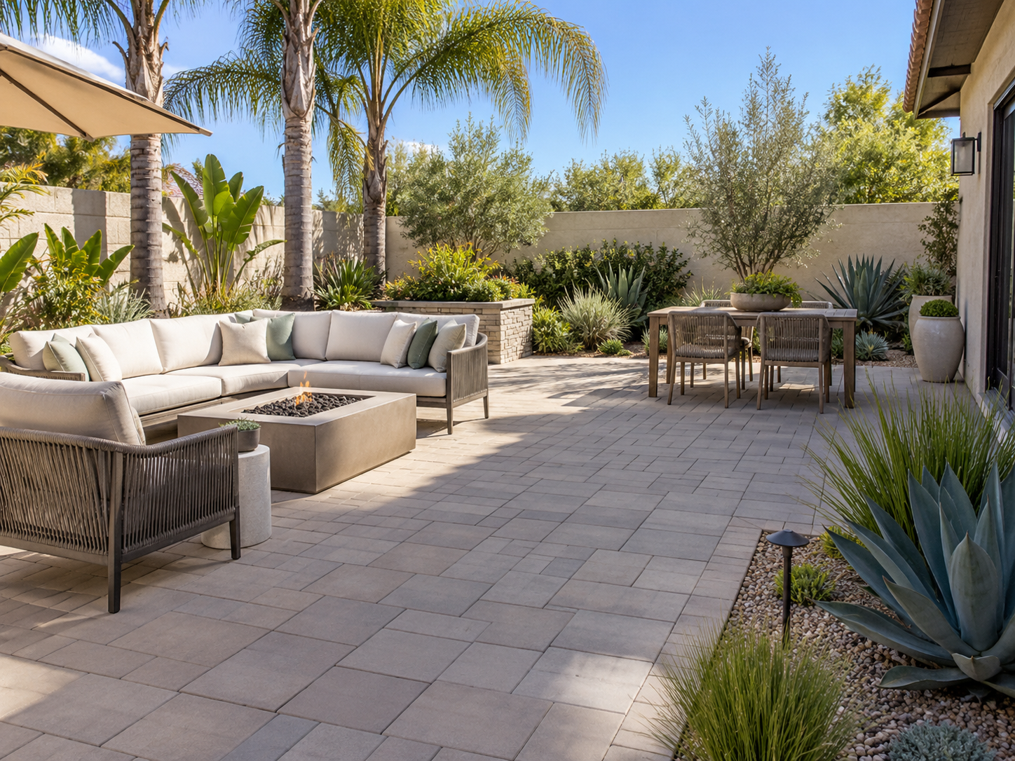 Spacious outdoor patio with beige pavers, a curved white sofa with multiple cushions, another seating chair, a rectangular fire pit, and a dining table with chairs, surrounded by lush green plants and tall palm trees under a clear blue sky.