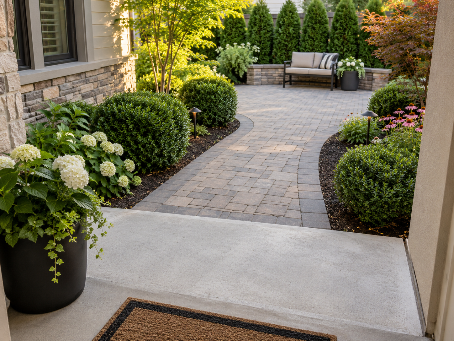 A garden pathway made of interlocking stone bricks with lush green bushes and flowers along the sides, leading to a patio area with a cushioned bench and decorative plants, surrounded by tall trees and a house wall.