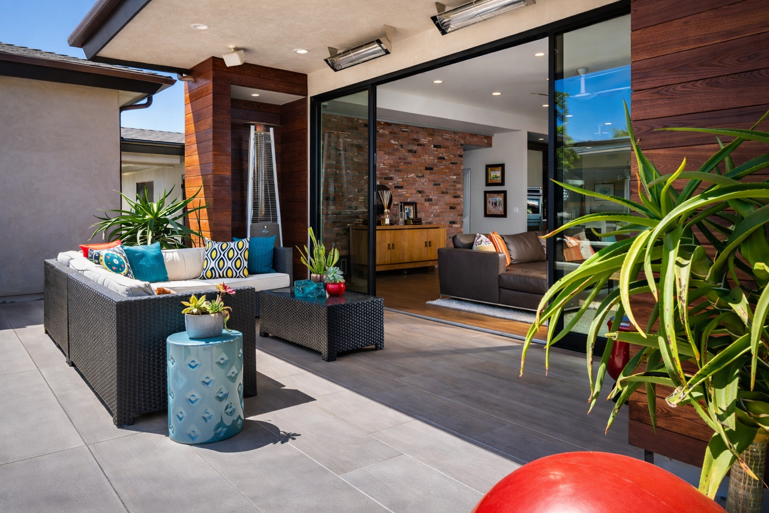 Outdoor patio with black wicker sectional sofa with colorful cushions, small blue side table, and potted plants. Inside, a living room visible through sliding glass doors has a brick accent wall, wooden sideboard, and brown leather couch.