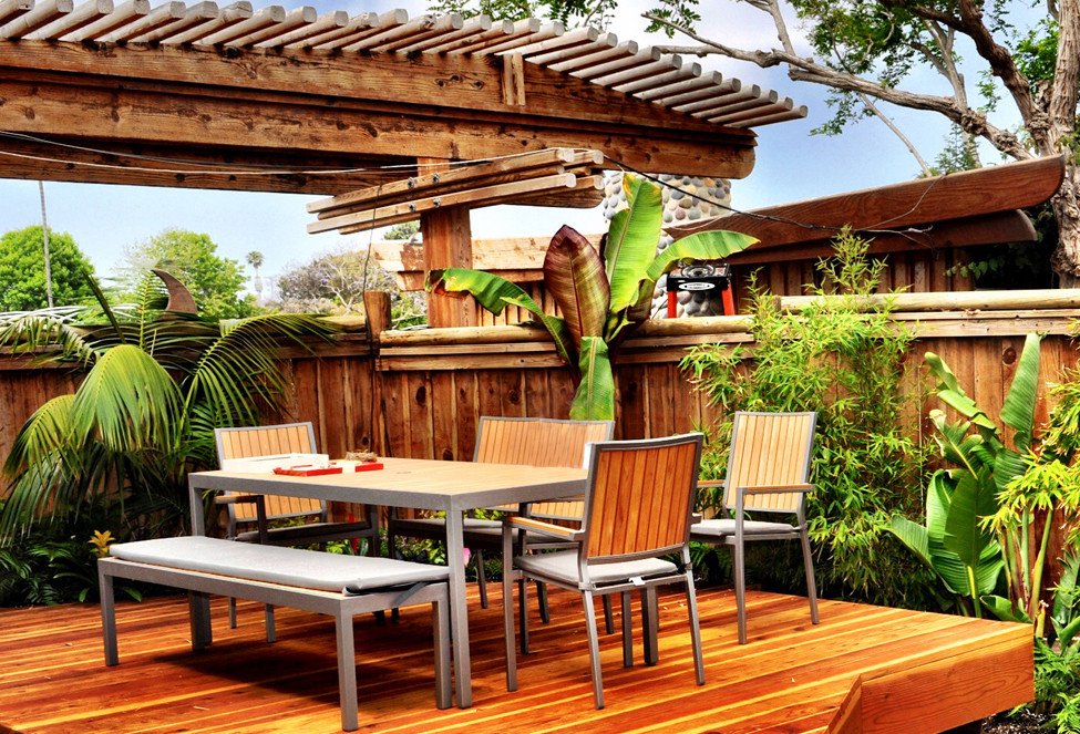 Outdoor patio with wooden deck, surrounded by a wooden fence and lush green plants, with a rectangular table and four chairs.