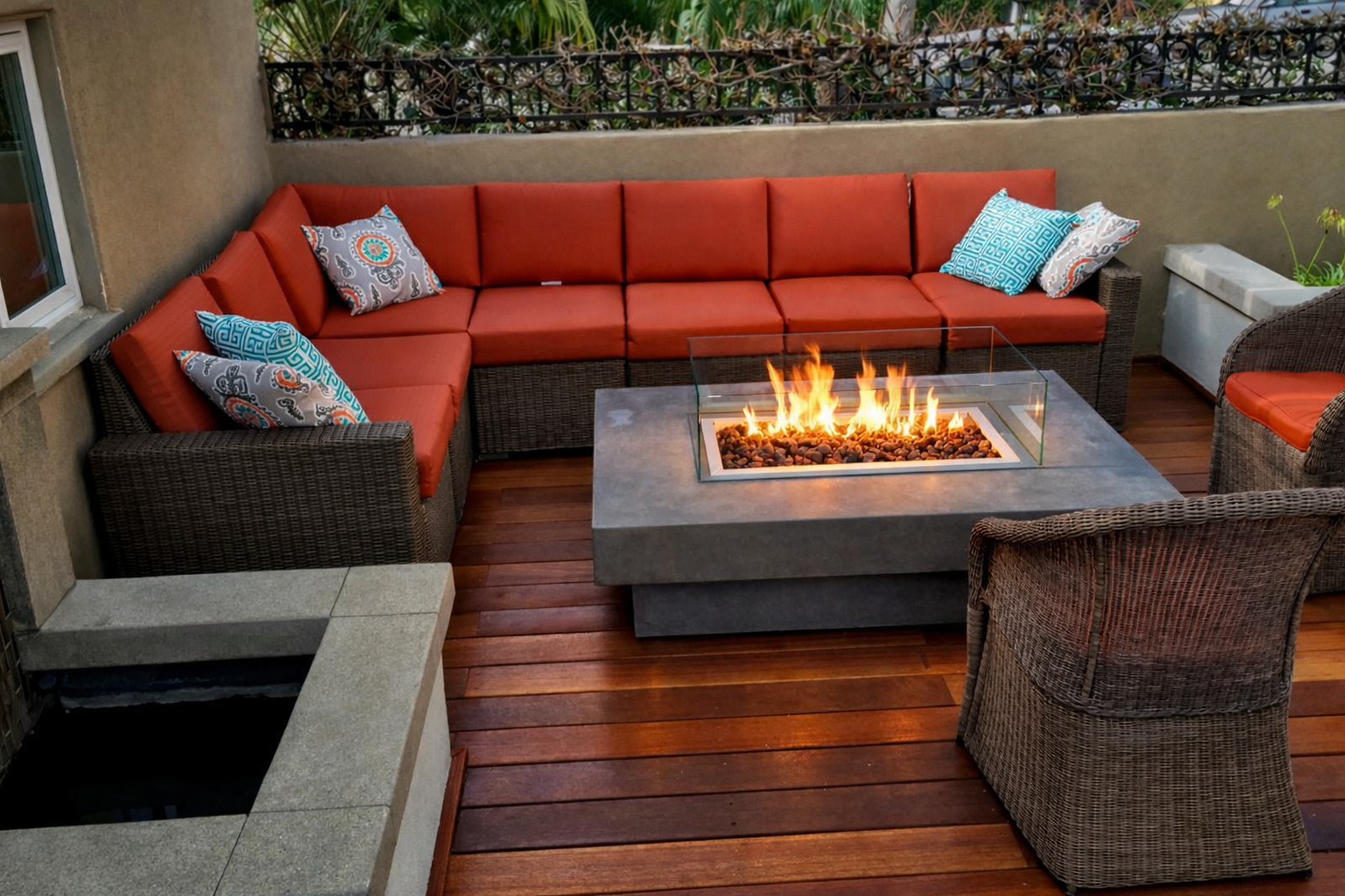 Outdoor patio with red cushioned wicker seating arranged around a modern rectangular fire pit on a wooden deck, with patterned pillows on the chairs and a garden fence in the background.  A contractor, who can design, build a fire pit near me. 