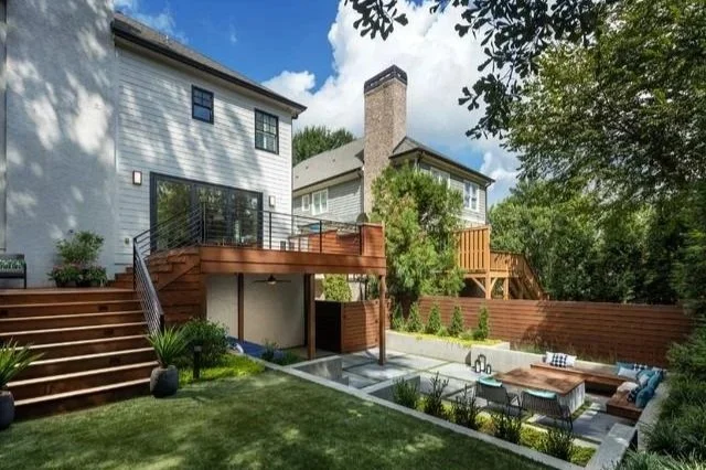 Backyard patio area with tiered wooden decks, outdoor furniture, and landscaped garden, attached to a white two-story house with a chimney and surrounding trees.
