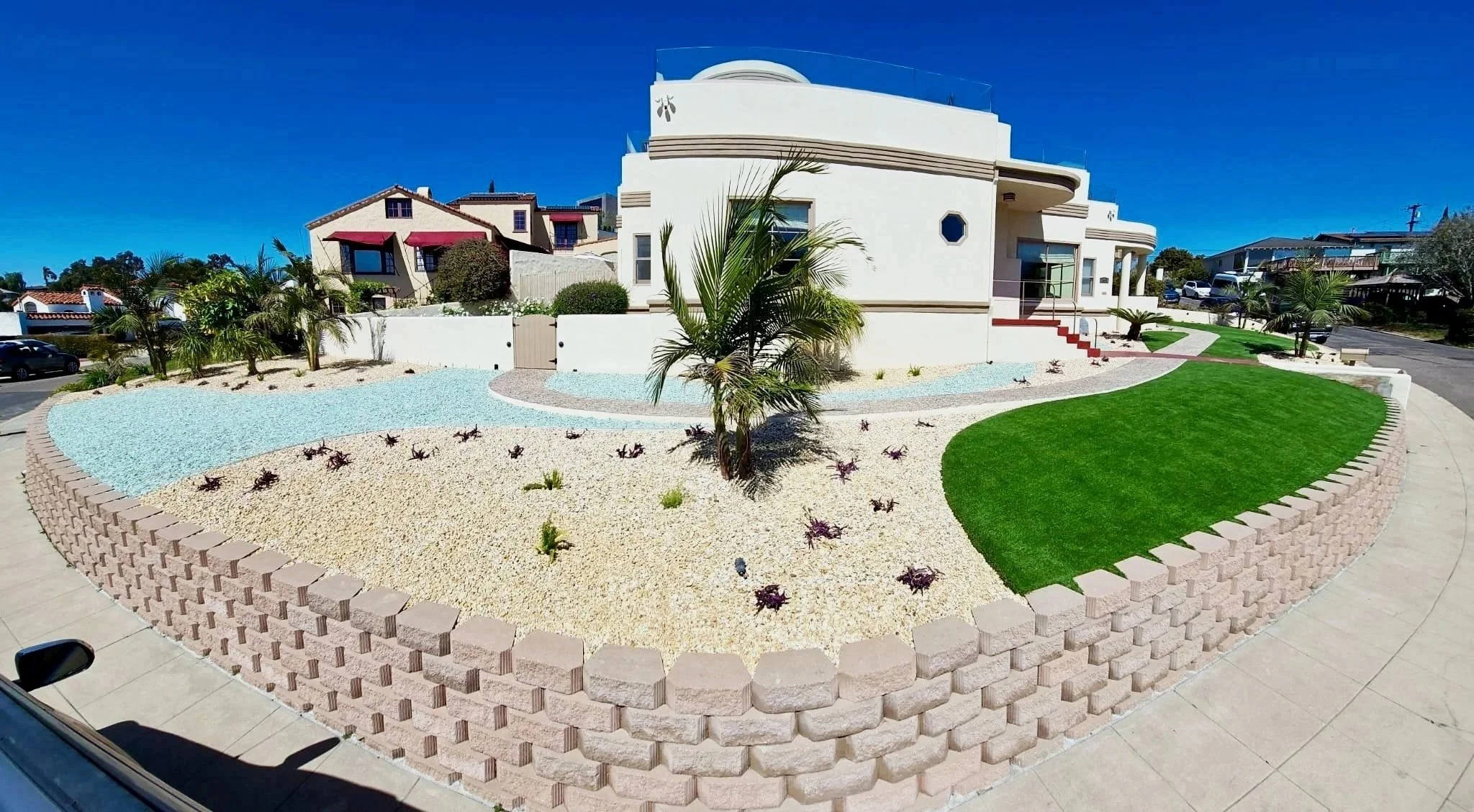 Modern house with a landscaped front yard featuring a curved green lawn, palm trees, and decorative rocks, surrounded by a low-key stone wall. Retaining walls contractor. They can build permitted walls near me in the San Diego area.